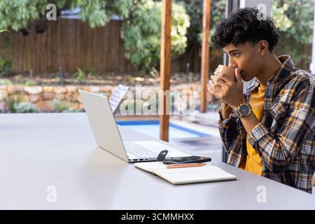 African american man sitting at patio table with coffee mug and using laptop, copy space Stock Photo