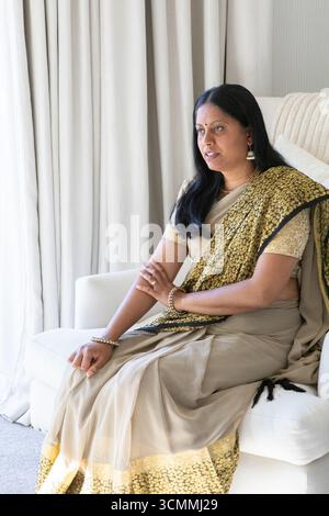 Senior indian woman sitting on upholstered armchair at home holding cushion wearing gold earring Stock Photo