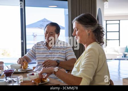 Senior couple sitting by wooden dining table at home sampling croissants, jam jar Stock Photo
