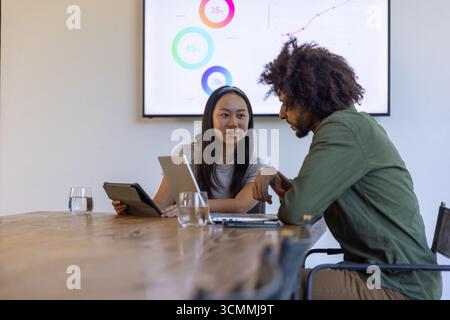 Diverse coworkers analyzing charts on screen while using tablet, laptop in meeting room at table Stock Photo