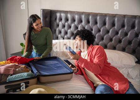 Diverse couple packing luggage on bed while checking passport on smartphone, placing straw hat Stock Photo