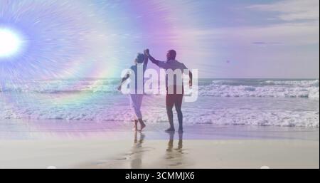 Dancing senior couple barefoot holding hands on beach shore, with breaking ocean waves, lens flare Stock Photo