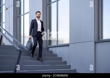 Confident businessman walking down stairs outside a modern office building, holding a briefcase and gazing ahead, exuding professionalism and ambition Stock Photo