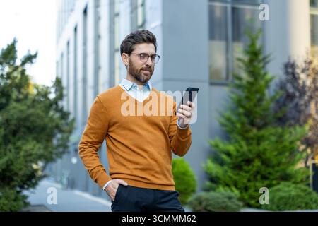 Business executive wearing glasses and an orange sweater, standing outdoors in a modern urban setting, uses a smartphone while holding one hand in his Stock Photo