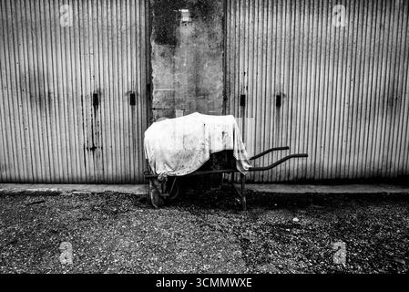 Black and white photo of an old abandoned wheelbarrow covered with cloth in front of a rusty garage door. Concept of decay, time passing, loneliness a Stock Photo