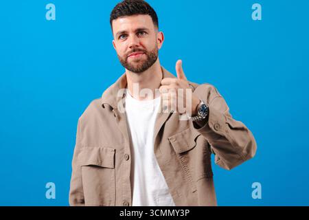 Young bearded man showing thumbs up gesture on blue background smiling Stock Photo