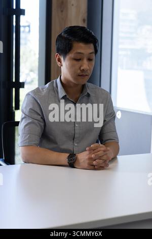 Asian man sitting at conference table by glass windows in mesh chair overlooking skyline Stock Photo