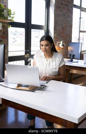 Woman typing on external keyboard at white table in office with laptop monitors and lamp Stock Photo
