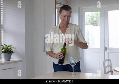 Woman opening green wine bottle at home kitchen island with potted plant and empty wine glass Stock Photo