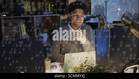 Developer wearing grey sweater analyzing binary code in office at night, with laptop and desk lamp Stock Photo