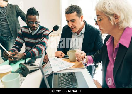 A diverse group of colleagues work together in a meeting, pointing at data charts and discussing business strategy while recording a corporate podcast Stock Photo