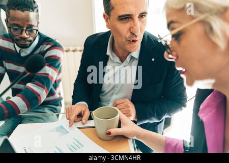 A diverse team of business colleagues intently analyze data charts and financial reports, collaborating closely during a strategic meeting or podcast Stock Photo