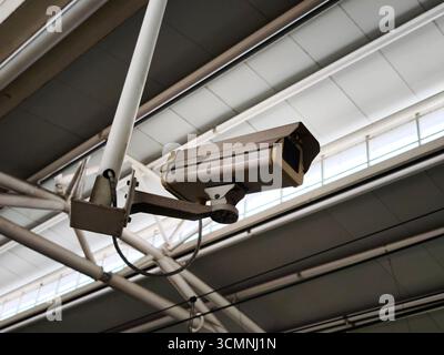 A security surveillance camera mounted on a metal frame inside a building. The camera is positioned under the ceiling structure, Stock Photo