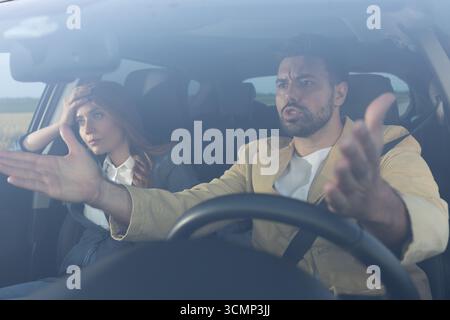 Angry driver on the road with arms open in outrage sitting next to stressed woman in passenger seat of car Stock Photo
