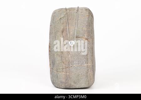 A silver necklace with a heart shaped pendant is displayed on a gray stone. The necklace is made of silver and has a diamond pendant. The stone is gra Stock Photo