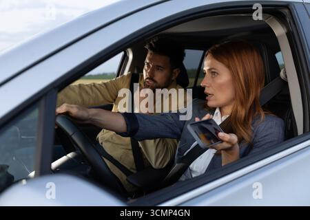 Couple in car debating directions, man pointing at road while woman drives with one hand holding onto smartphone Stock Photo