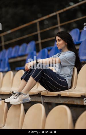 A brunette girl sits alone in the empty stadium stands. She wears a blue T shirt and casual pants enjoying the quiet atmosphere in this spacious venue. Stock Photo