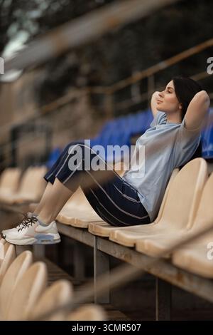 Brunette girl sits comfortably alone in the empty stadium stands wearing a blue T shirt and casual pants. She appears serene soaking in the atmosphere of the venue. Stock Photo
