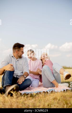 A family sits on a blanket in a grassy field under a clear blue sky, sharing joyful moments as they laugh and bond together. The atmosphere is warm an Stock Photo