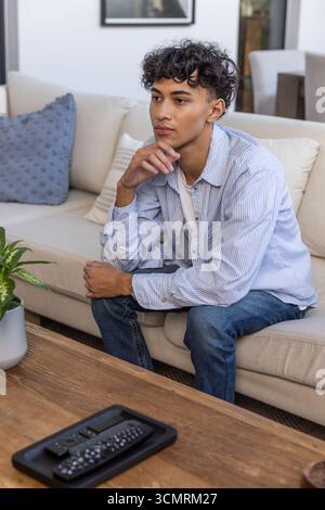 Man in his 20s sitting on sofa at home, remote controls with potted plant nearby Stock Photo