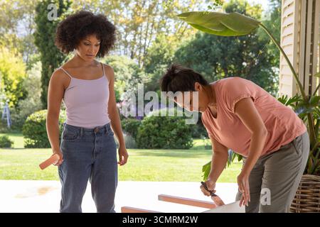Diverse mother and daughter painting wooden planks on porch with pink paint and brushes Stock Photo