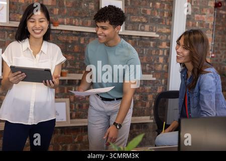 Diverse coworkers collaborating on project at desk in office with tablet, papers, pencil, laptop Stock Photo