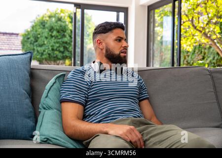 Man wearing striped t-shirt sitting on sofa at home with windows, teal cushions and headphones Stock Photo
