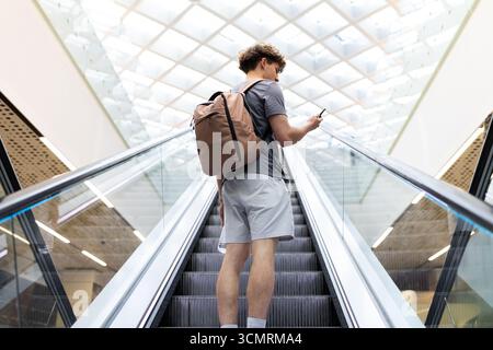 Young Caucasian man with backpack using smartphone while standing on airport escalator Stock Photo