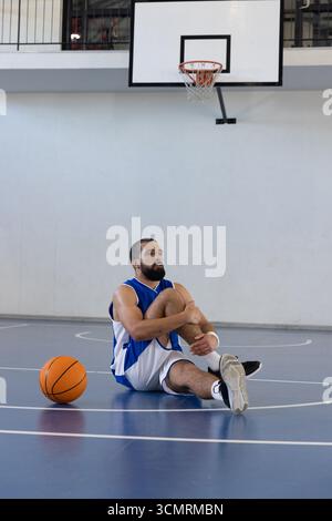 Male basketball player in jersey holding knee sitting on court near basketball, copy space Stock Photo