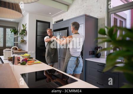 Senior couple wearing aprons dancing and holding hands in kitchen by island with vegetables Stock Photo