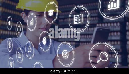 Examining smartphone female technician wearing safety helmet and shirt in depot with floating icons Stock Photo