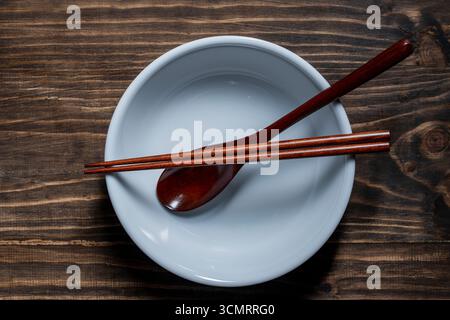 A pair of wooden chopsticks and spoon lying across an empty white soup bowl on wooden table, close up, top view Stock Photo