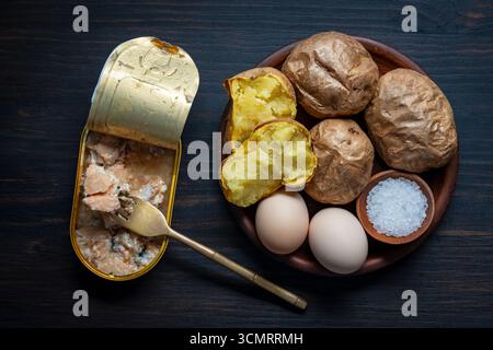 Open tin can of salmon fish fillet and baked potatoes whole in their skins, soft-boiled eggs, salt in clay plate on wooden table, close up, top view. Stock Photo