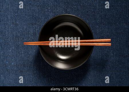 A pair of wooden chopsticks lying across an empty black soup bowl on blue fabric background, close up, top view Stock Photo