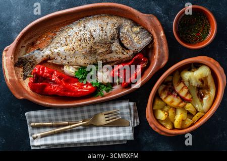 Delicious baked dorado fish with spices in a clay plate and fried vegetables, close up, top view Stock Photo