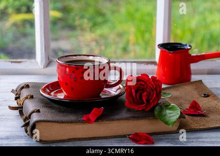 Ceramic red cup of coffee, red rose and notebook on the table in front of the window overlooking the summer garden, close up Stock Photo