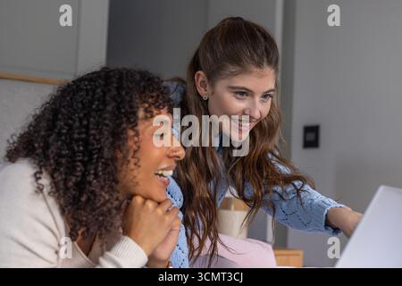 Diverse female colleagues leaning over desk, smiling, reviewing content on laptop in home office Stock Photo