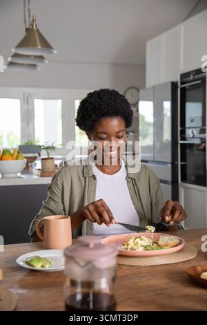 African american woman cutting into avocado toast on pink plate in kitchen with coffee carafe Stock Photo