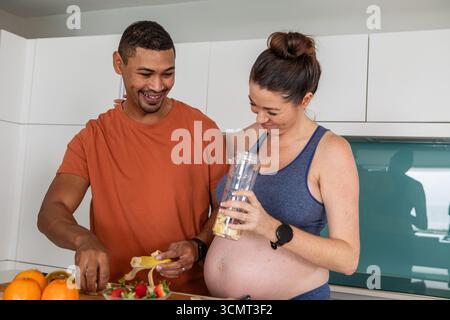 Diverse couple slicing banana, strawberries on board for smoothie in kitchen holding blender cup Stock Photo