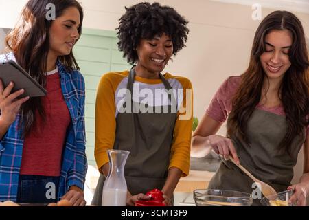 Diverse female friends standing at kitchen island preparing recipe using tablet, mixing bowl Stock Photo