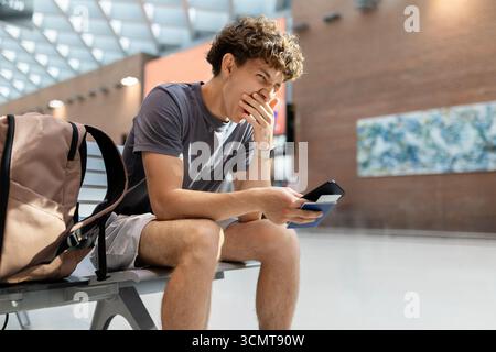 Young man sitting at airport bench yawning tired with passport and smartphone Stock Photo