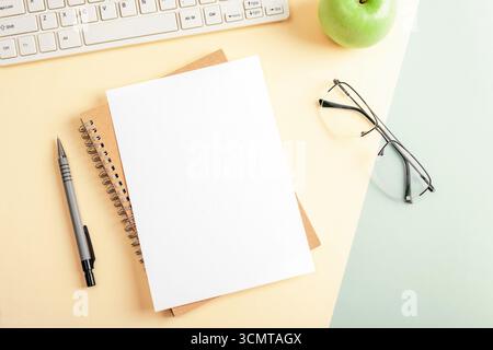 White blank paper on spiral notebook with pen, computer keyboard, apple and glasses on yellow and green desk. Top view, flat lay, mockup. Stock Photo