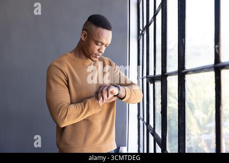 African american man wearing tan sweatshirt standing by window in loft checking smartwatch Stock Photo