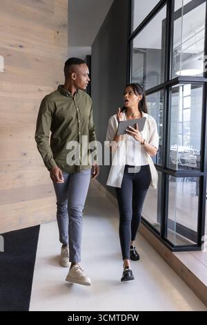 Coworkers walking through office corridor beside wood-panel, glass partitions holding tablet Stock Photo
