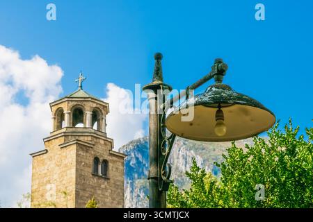 Rustic street lamp stands near a historic stone church tower with cross on top, captured under bright daylight with blue sky and mountain in the backg Stock Photo
