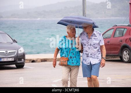 Vodice, Croatia - August 21, 2025: Senior couple walking under umbrella in a parking area on a rainy summer day on Croatian coast Stock Photo