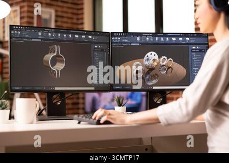 Female engineer reviewing mechanical clamp and gear prototype on dual monitors using CAD software. Creative asian woman working from home workstation, creating 3d industrial tools for construction. Stock Photo