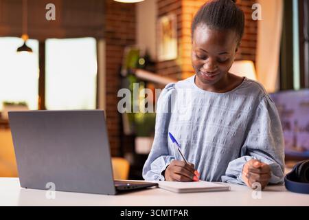 Focused female freelancer working indoors, reviewing sales performance on laptop and taking notes. Remote worker writing ideas in notepad while managing reporting and organizational tasks. Stock Photo