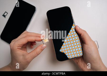 High-angle view of woman inserting sim card into new smartphone, with another phone and sim ejector tool nearby on white table, during mobile device Stock Photo