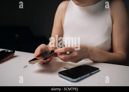 Close-up cropped shot of woman using sim ejector tool to open sim card tray of mobile phone, sitting at white table, engaged in process of managing Stock Photo